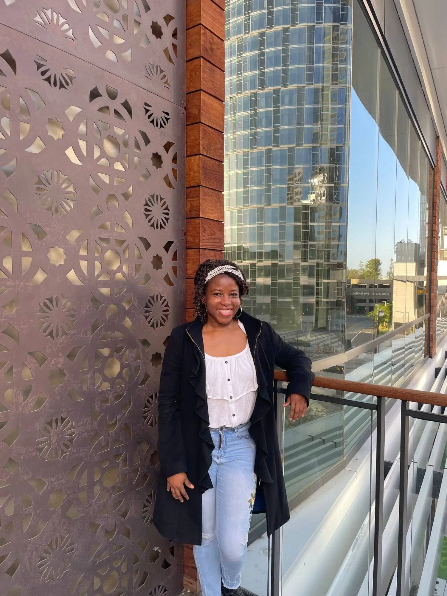 Young woman in black coat and light jeans leans on balcony, glass building and panel behind her.