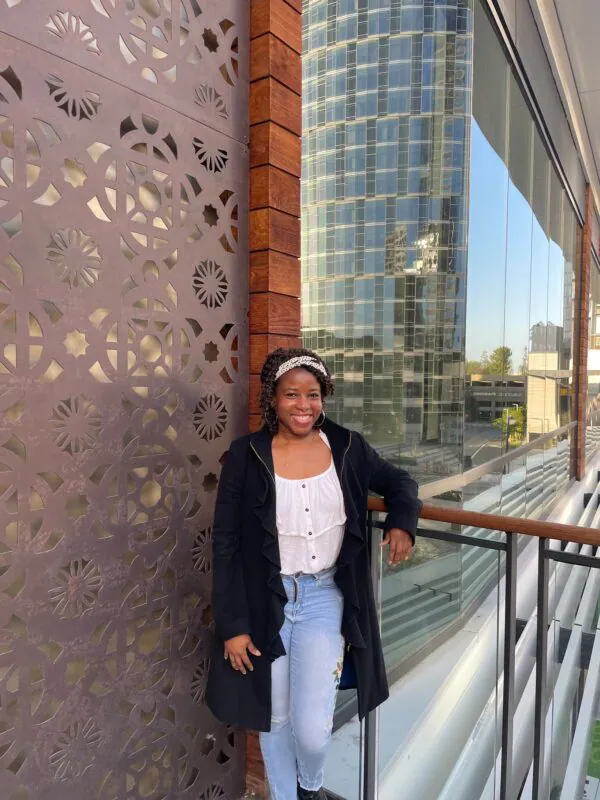 Smiling woman in casual outfit leaning on balcony railing, decorative wall, skyscraper backdrop, clear blue sky.